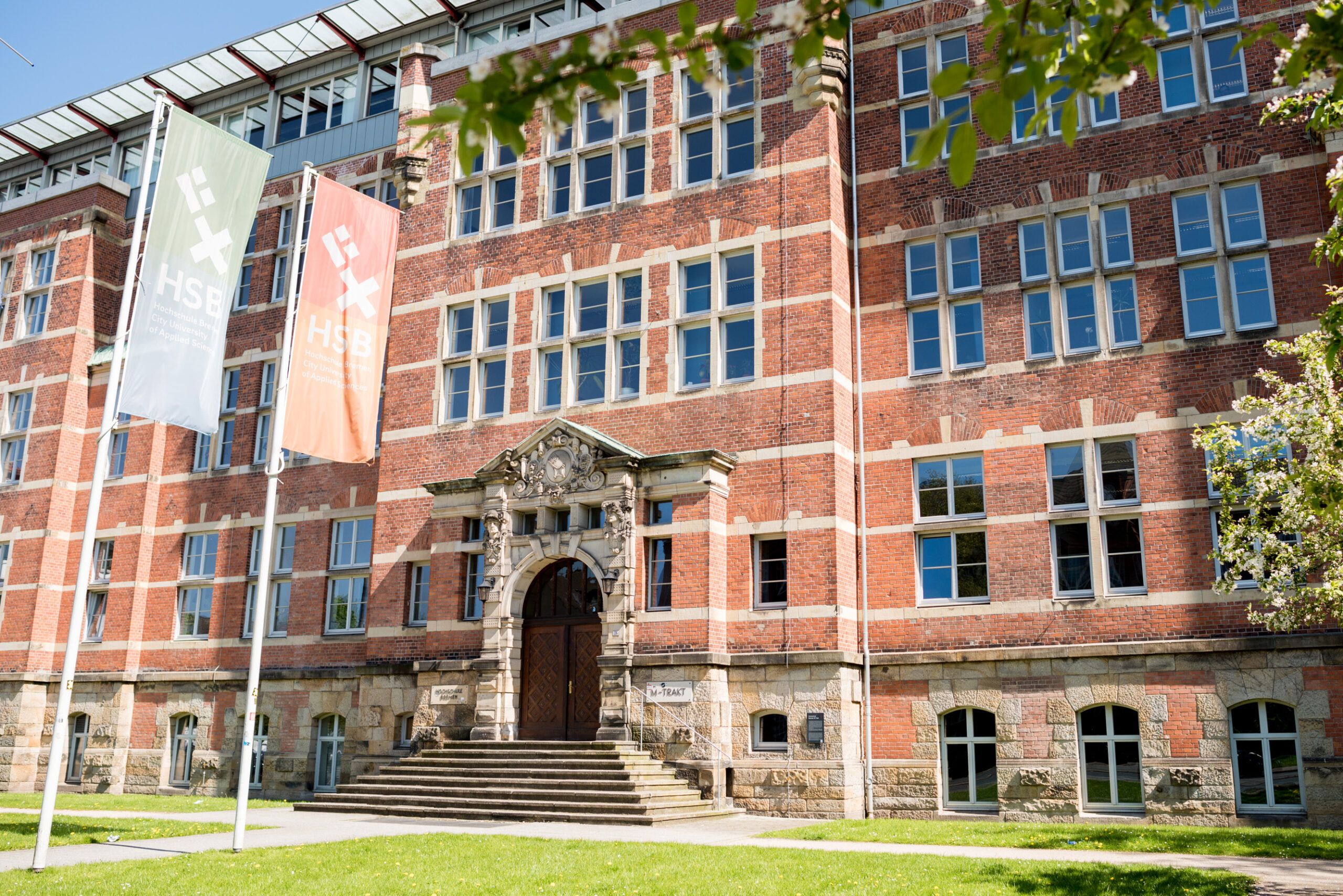 Der Eingang des M-Gebäude der Hochschule Bremen mit zwei Flaggen davor. Auf beiden Flaggen ist das Logo der Hochschule Bremen.