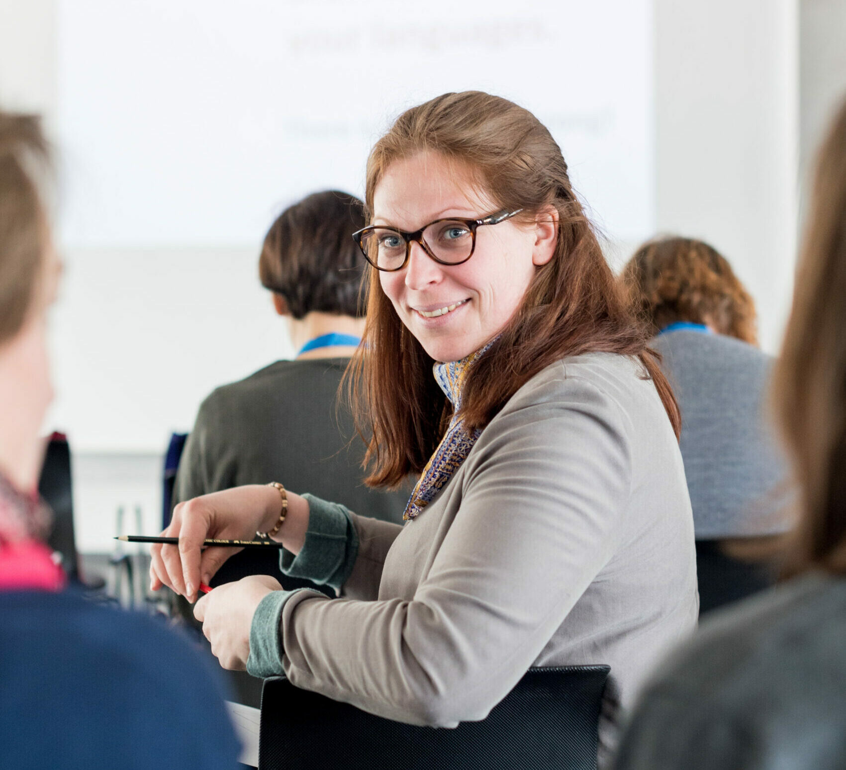 Ein Frau sitzt in einer Stuhlreihe bei einem Vortrag und dreht sich freundlich zu den Menschen um, die hinter ihr sitzen.