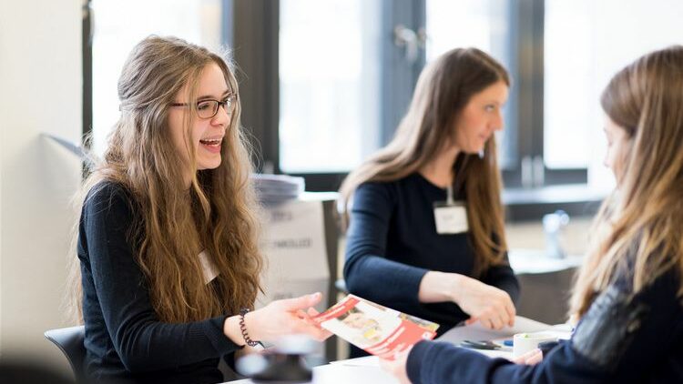 Zwei Personen unterhalten sich miteinander. Eine Person reicht der anderen Person einen Flyer herüber.