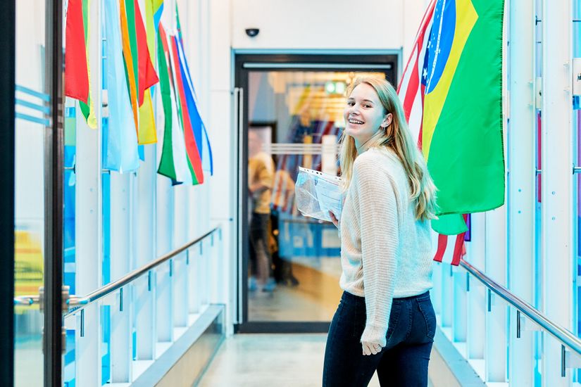 Eine junge Frau steht im blauen Gang der Hochschule Bremen und lächelt in die Kamera. Links und rechts von ihr sind diverse Länderflaggen.
