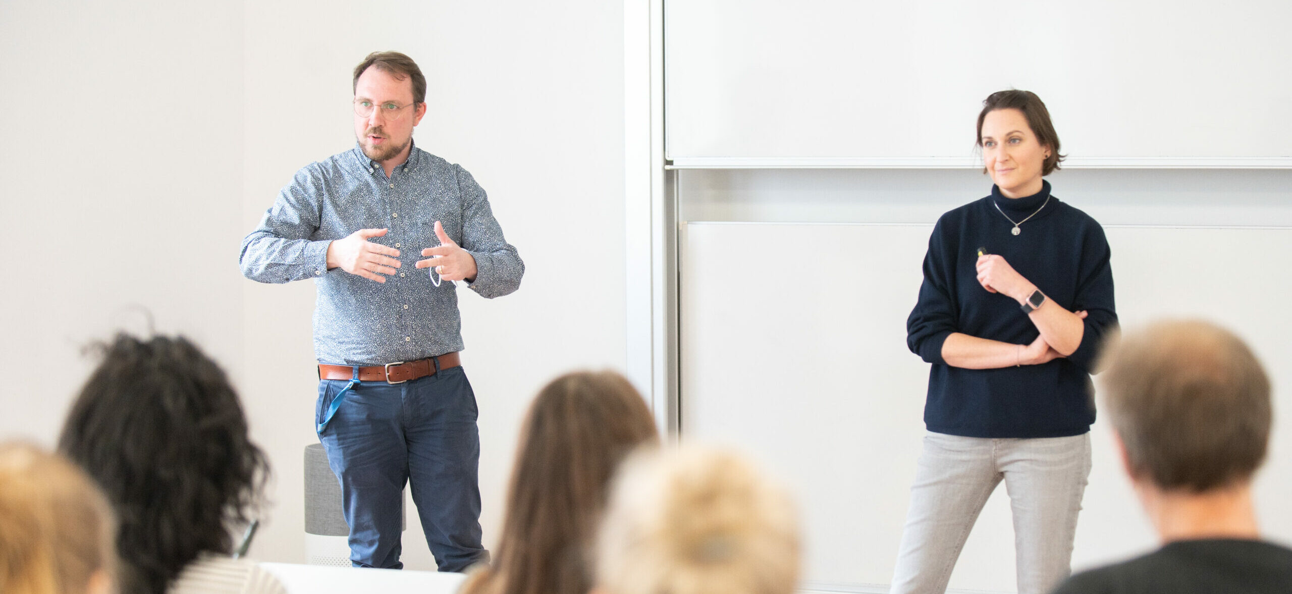 Zwei Lehrpersonen stehen vor einer Gruppe Studierender im Vorlesungsraum und erklären etwas.