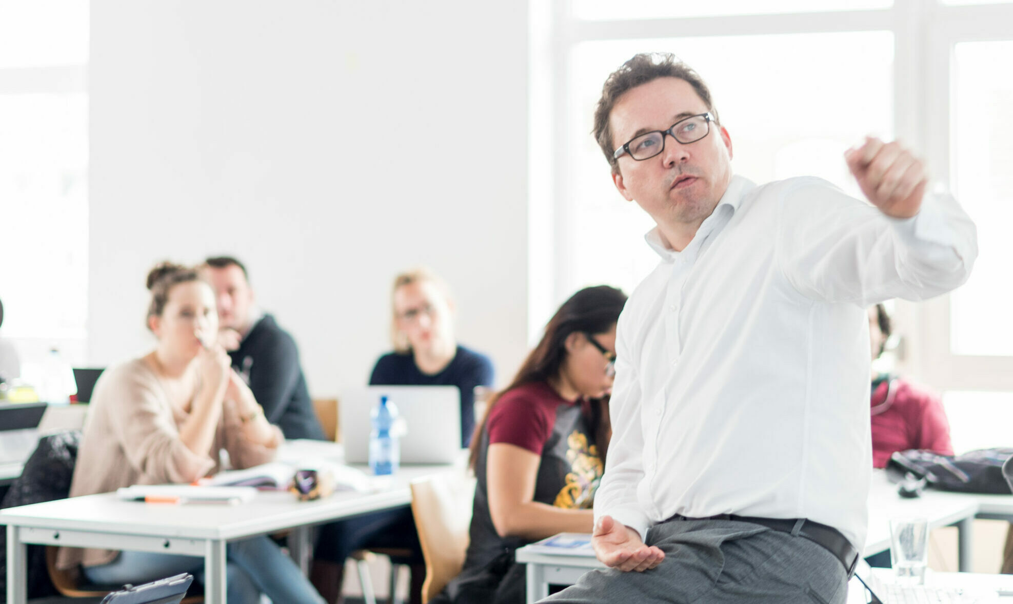 Ein Dozent sitzt vor einer Gruppe Studierender auf einem Tisch und erklärt etwas auf der Tafel hinter ihm.