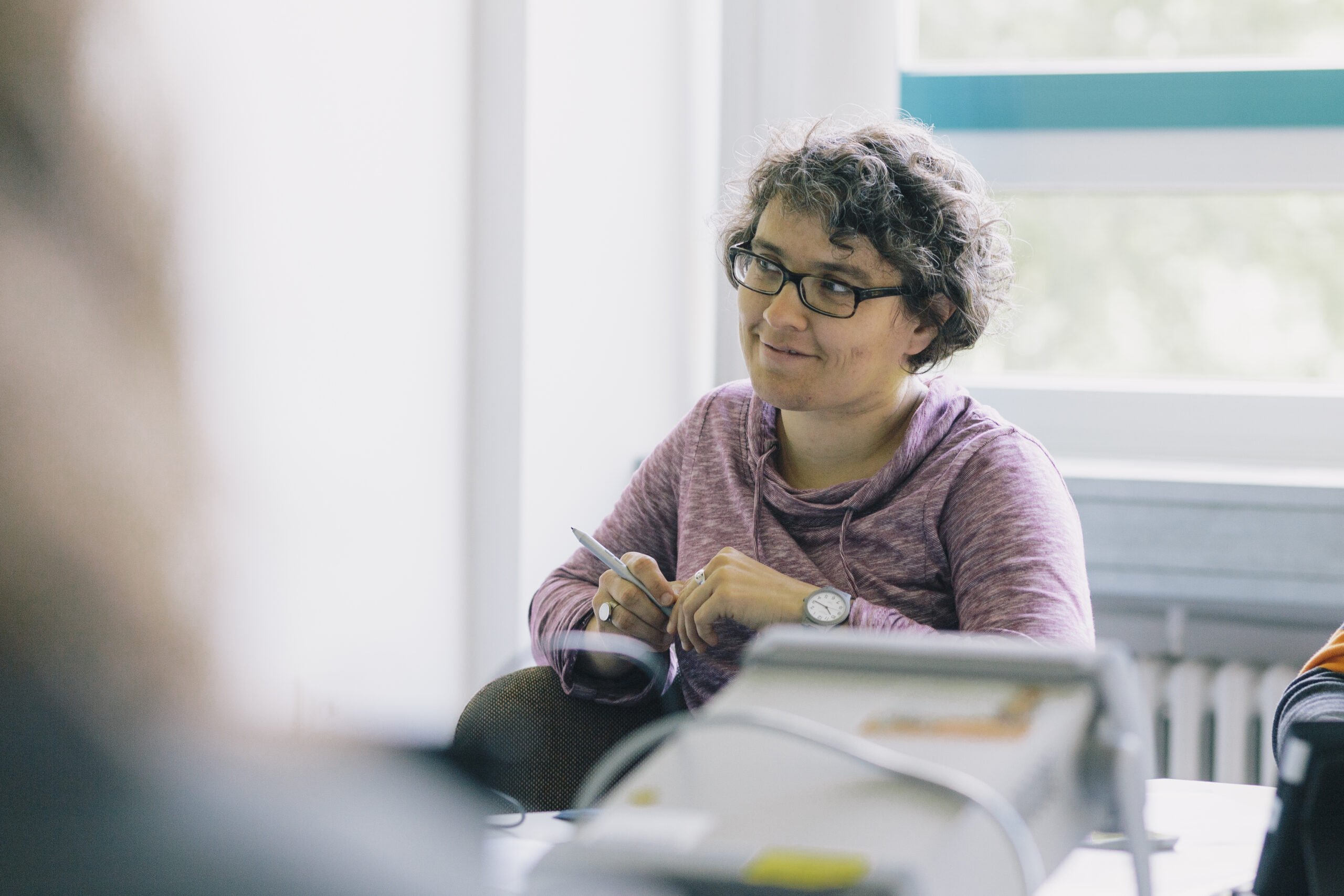 Eine Person sitzt in einem Veranstaltungsraum mit einem Stift in der Hand und hört jemandem zu.