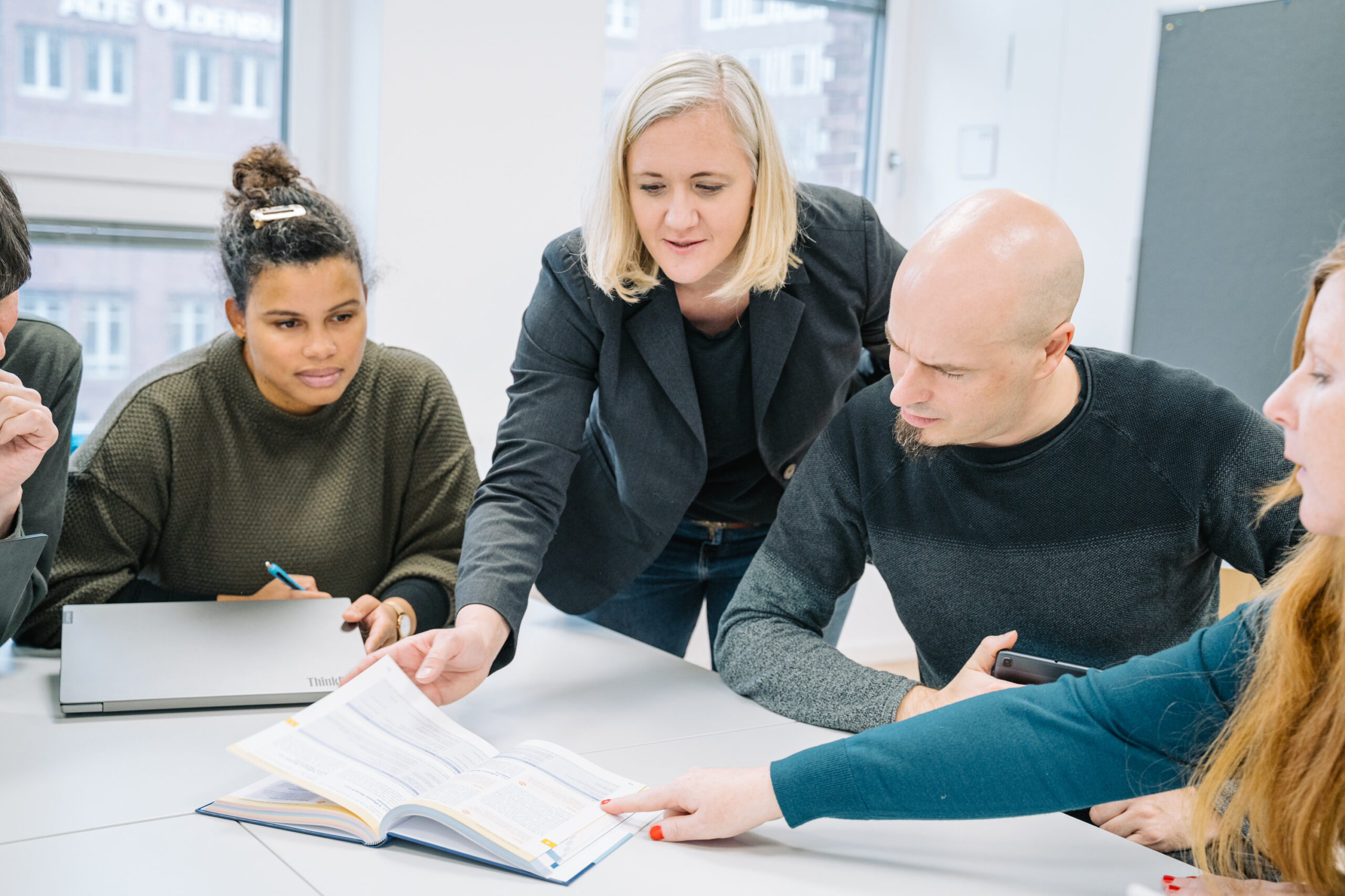 Eine Dozentin erklärt einer Arbeitsgruppe etwas in einem Buch.