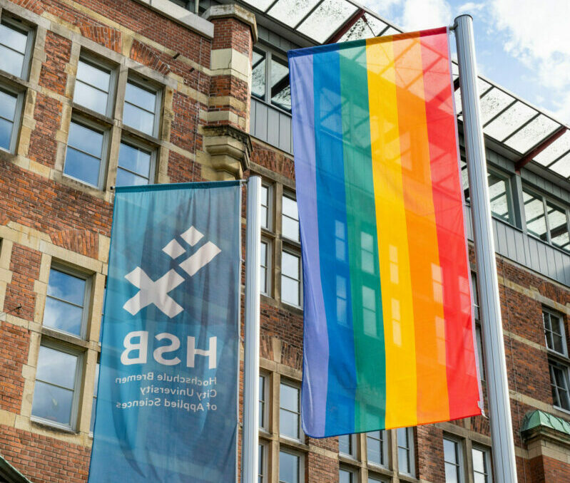 Zwei gehisste Flaggen vor dem M-Gebäude der Hochschule Bremen. Die eine Flagge zeigt das Logo der H S B, die andere Flagge ist die Regenbogenfahne mit den Farben lila, blau, grün, gelb, orange und rot als Symbol der L G B T Q Bewegung.