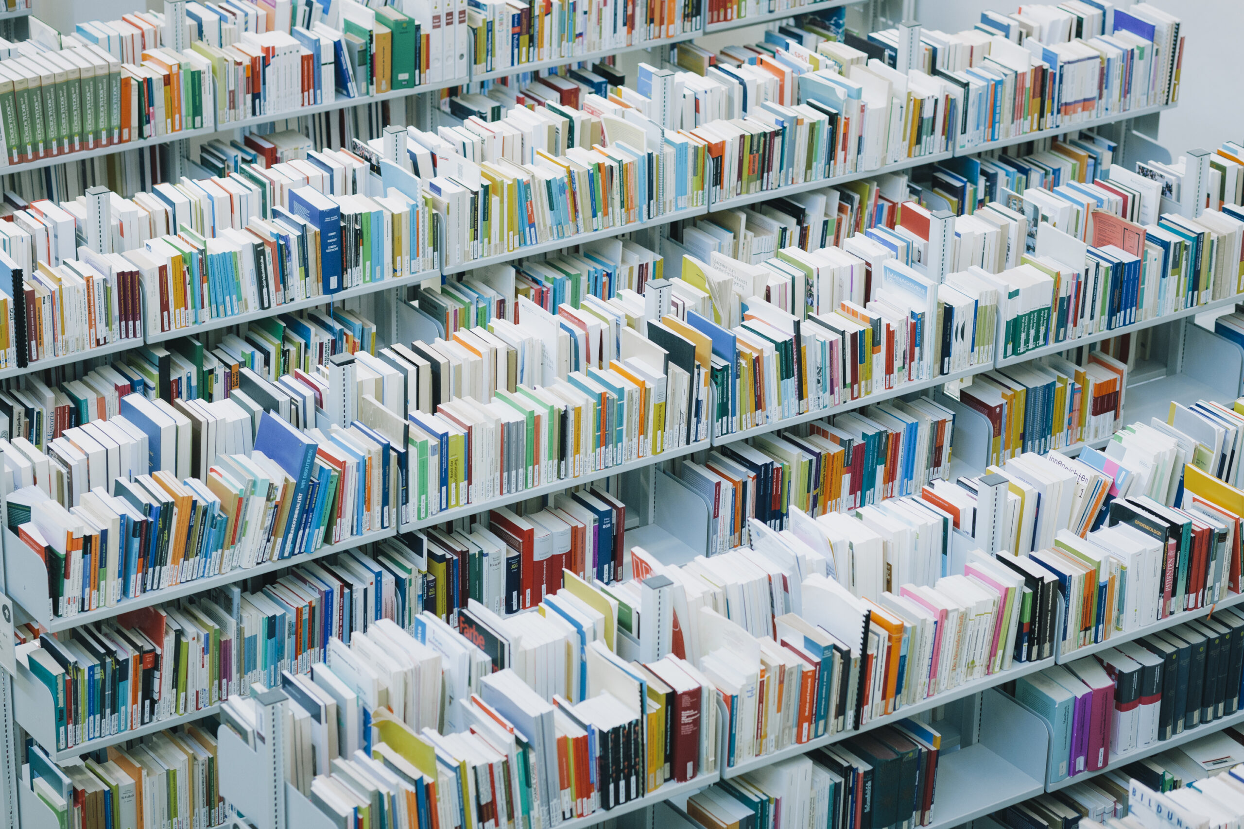 Zu sehen sind mehrere weiße, mehrstöckige Bibliotheksregale mit unzähligen Büchern verschiedener Größe und Farbe.